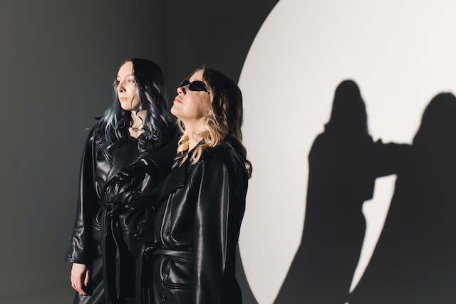 Two girls in black leather coats. They're serious and one of them is wearing black sunglasses. We can see their shadow on the wall behind them. 