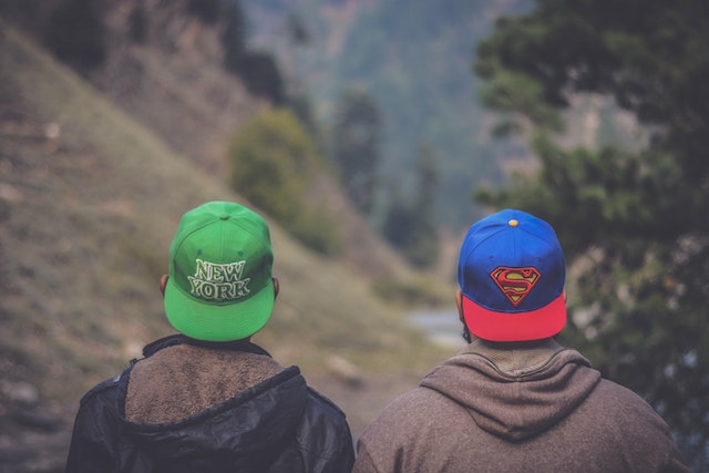 Two men sitting in the woods looking in the distance. We can see their back. They both wear caps and hoodies. The hoodies are black and brown. The first one's cap is green and it says "New York". The second one's cap is blue and red with the Superman's sign on it.