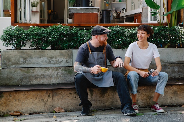 A man and a woman sitting at the bench and talking. He's wearing a cap and an apron while holding a cup of coffee. She's wearing jeans and a white T-shirt. They're smiling.